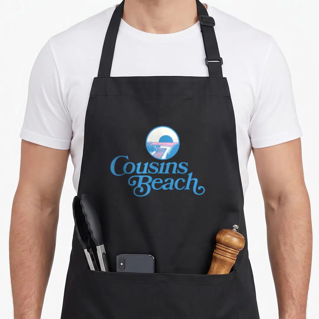 Cousin pretty apron - Retro circular beach scene with blue script reading Cousins Beach — shown on a black cotton canvas apron against a white background