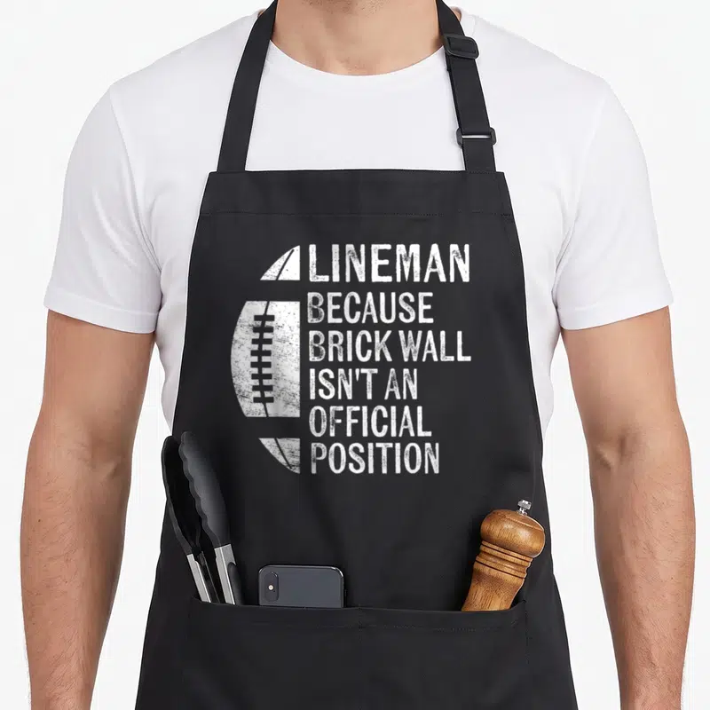 Wall football apron - Distressed white football next to stacked text reading Lineman Because Brick Wall Isn't — shown on a black cotton canvas apron against a white background