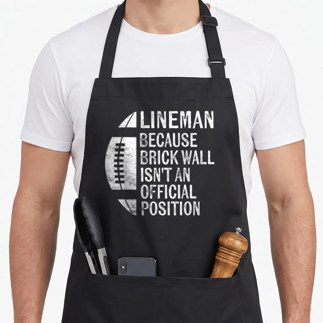 Wall football apron - Distressed white football next to stacked text reading Lineman Because Brick Wall Isn't — shown on a black cotton canvas apron against a white background