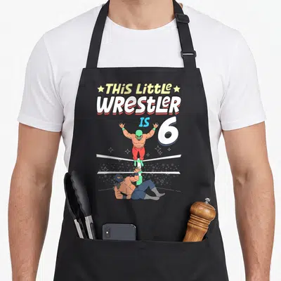 Wrestling 6th birthday apron - This Little Wrestler Is 6 text with green masked wrestler jumping over blue masked — shown on a black cotton canvas apron against a white background