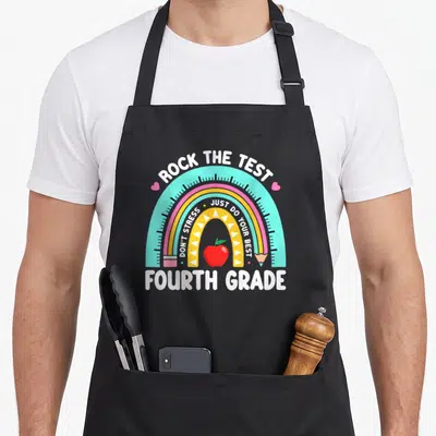Test 4th teacher apron - Rainbow with rock the test text, apple, pencil, and fourth grade label — shown on a black cotton canvas apron against a white background