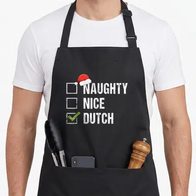 Dutch christmas santa apron - Checklist design with NAUGHTY NICE DUTCH text, red Santa hat, and green checkmark — shown on a black cotton canvas apron against a white background