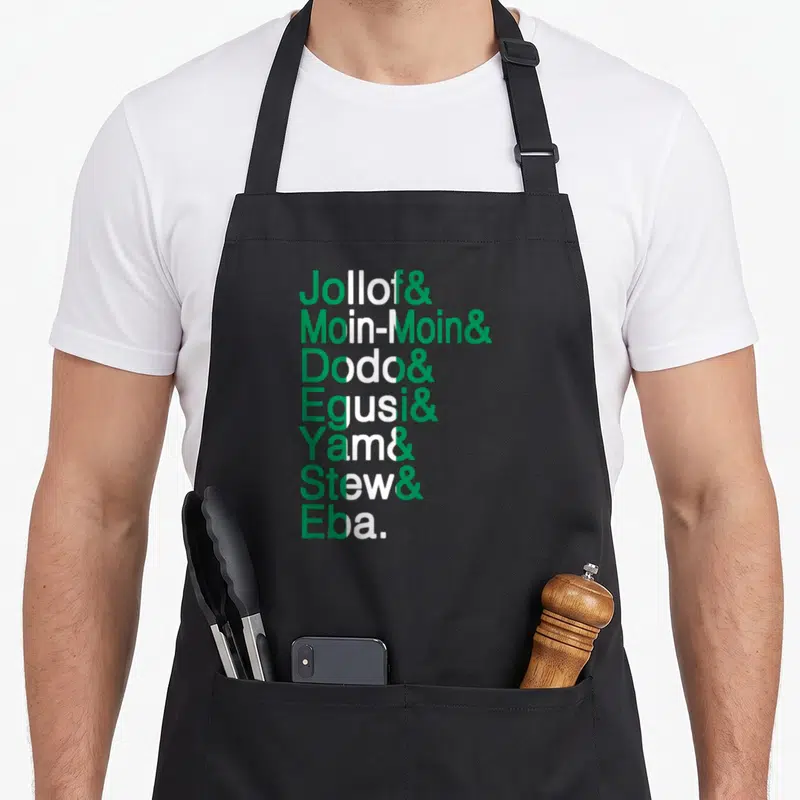 Nigerian food aprons - Green and white stacked text listing Jollof Moin-Moin Dodo Egusi Yam Stew Eba with central stripe — shown on a black cotton canvas apron against a white background