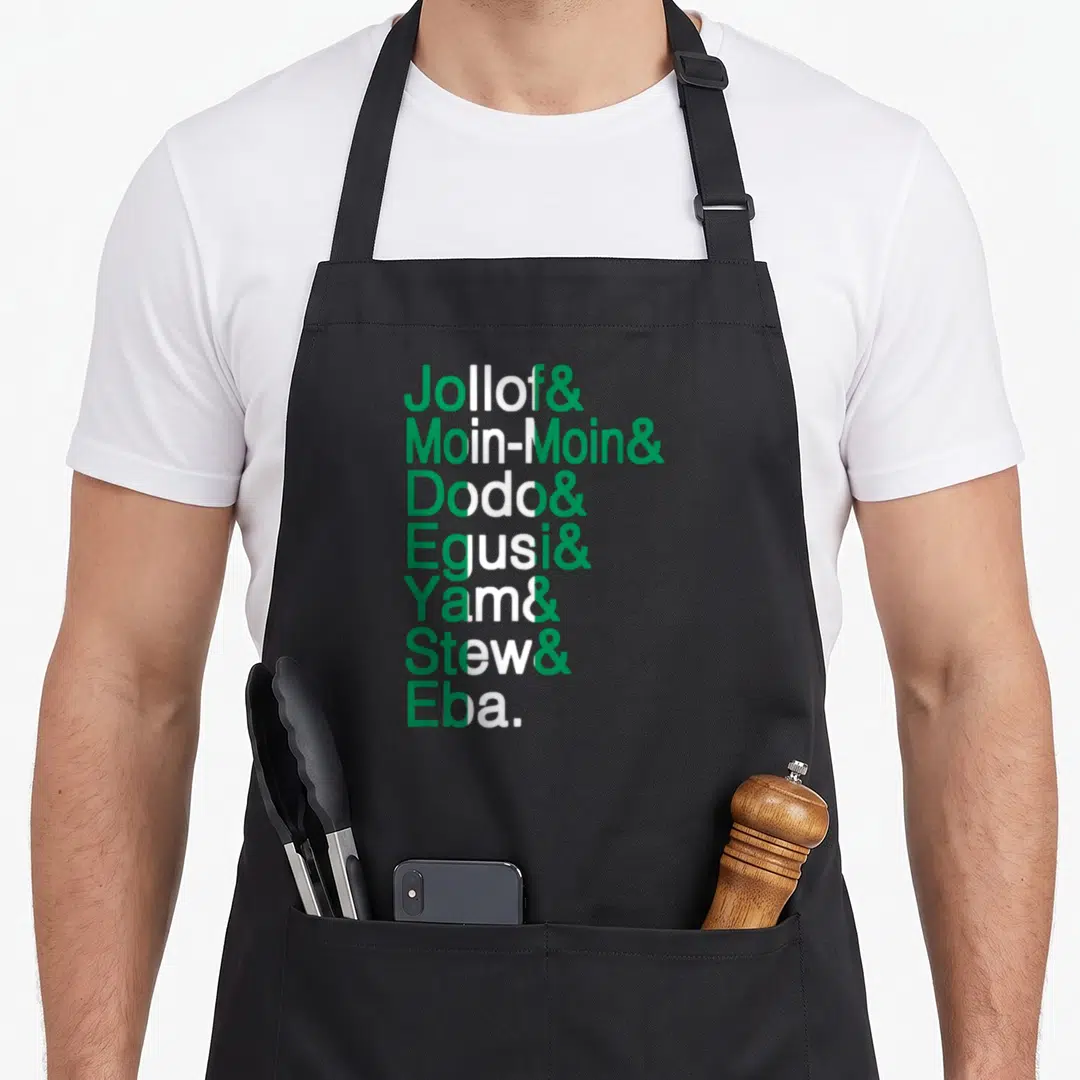 Nigerian food aprons - Green and white stacked text listing Jollof Moin-Moin Dodo Egusi Yam Stew Eba with central stripe — shown on a black cotton canvas apron against a white background