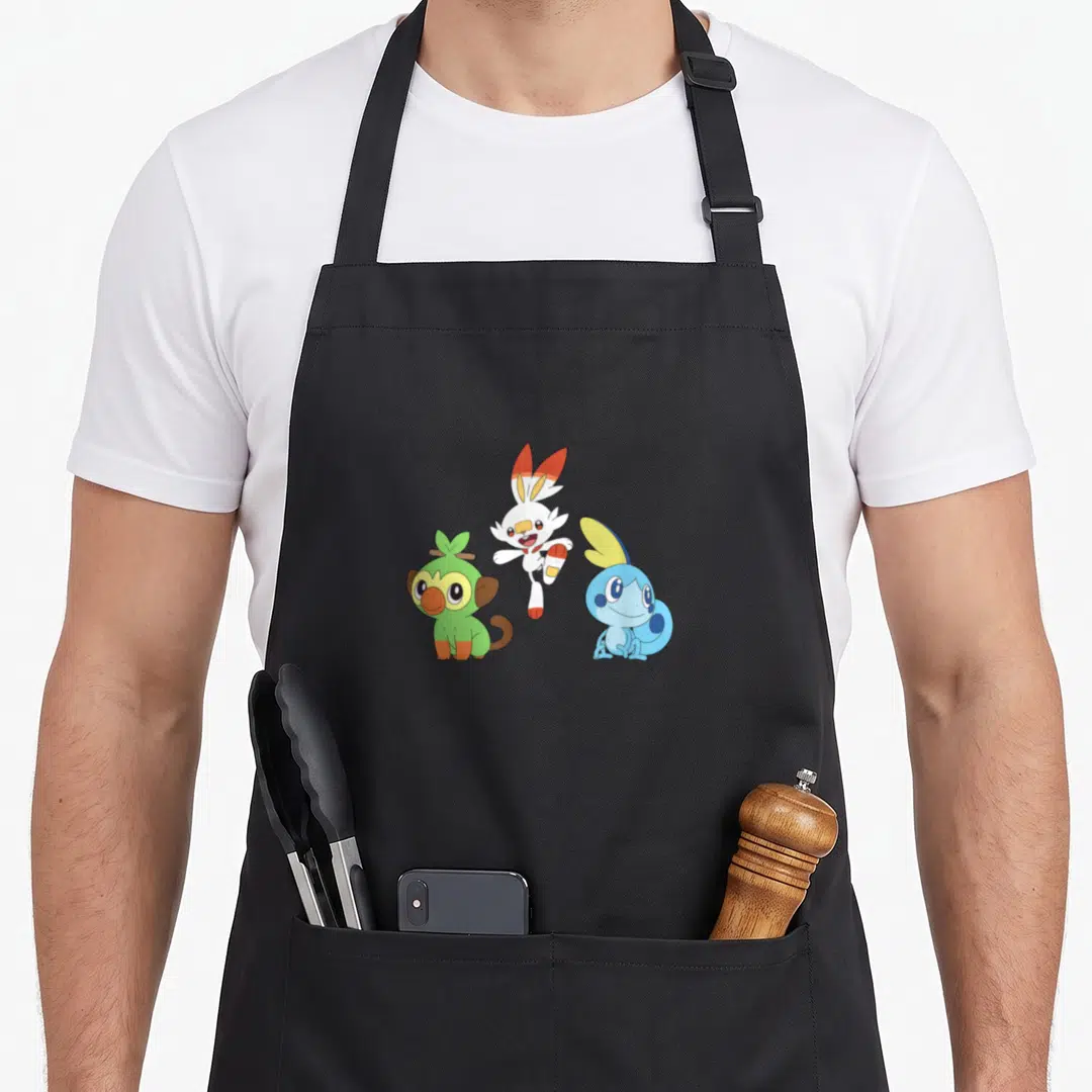 Scorbunny trend apron - Three Pokémon Grookey Scorbunny Sobble cartoon cluster green white blue orange — shown on a black cotton canvas apron against a white background
