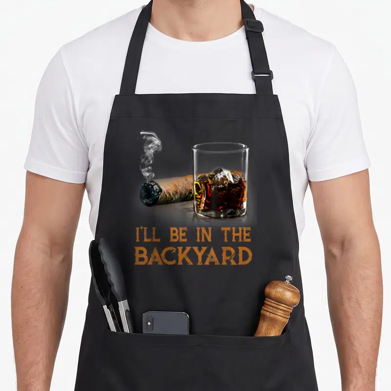 Ill bourbon apron - Smoking cigar and whiskey glass with orange text I'll Be In The Backyard — shown on a black cotton canvas apron against a white background