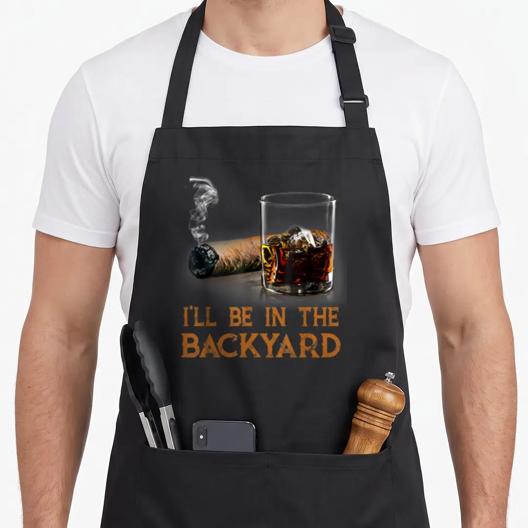 Ill bourbon apron - Smoking cigar and whiskey glass with orange text I'll Be In The Backyard — shown on a black cotton canvas apron against a white background