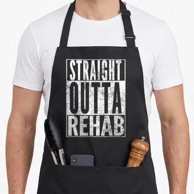 Straight addiction recovery apron - White stacked text reading STRAIGHT OUTTA REHAB inside a rectangular frame — shown on a black cotton canvas apron against a white background