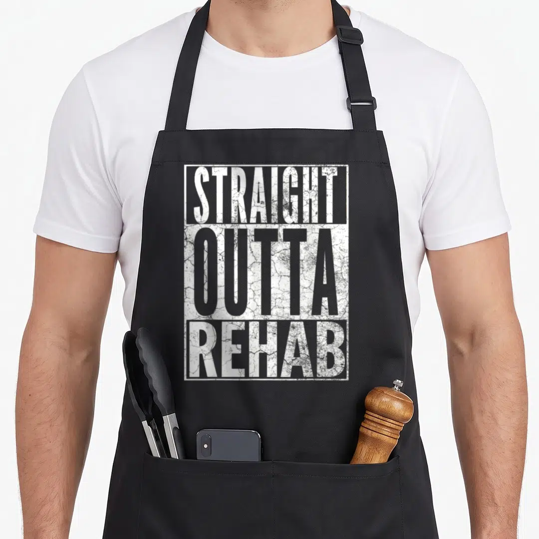 Straight addiction recovery apron - White stacked text reading STRAIGHT OUTTA REHAB inside a rectangular frame — shown on a black cotton canvas apron against a white background