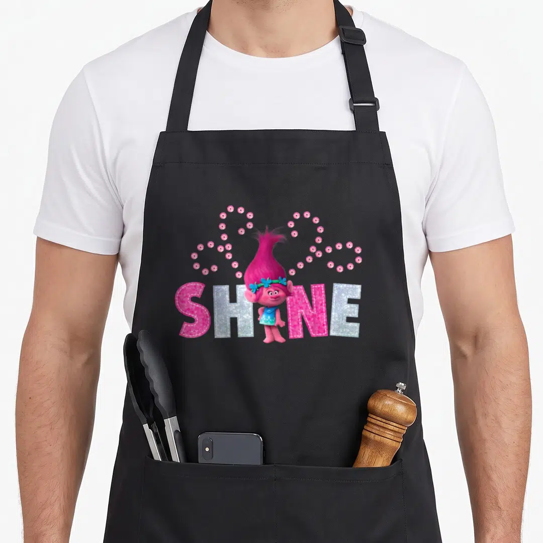 Poppy troll apron - Pink Poppy character standing before silver Shine text with pink gem heart arrangement — shown on a black cotton canvas apron against a white background