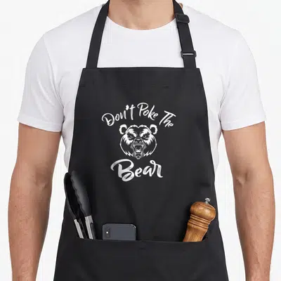 Grumpy mama apron - White cartoon bear head with arched text Don't Poke The Bear — shown on a black cotton canvas apron against a white background