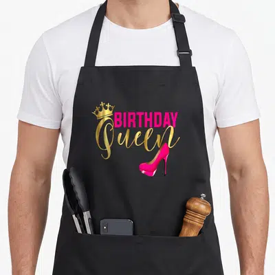 Birthday queen of baking apron - Pink BIRTHDAY text, gold Queen script, crown, and high heel shoe — shown on a black cotton canvas apron against a white background