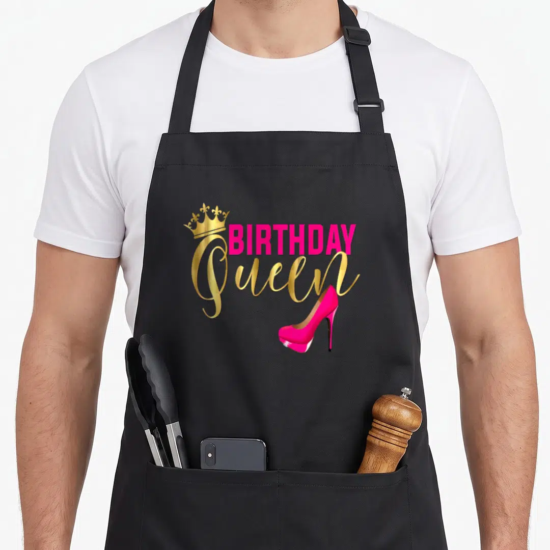 Birthday queen of baking apron - Pink BIRTHDAY text, gold Queen script, crown, and high heel shoe — shown on a black cotton canvas apron against a white background