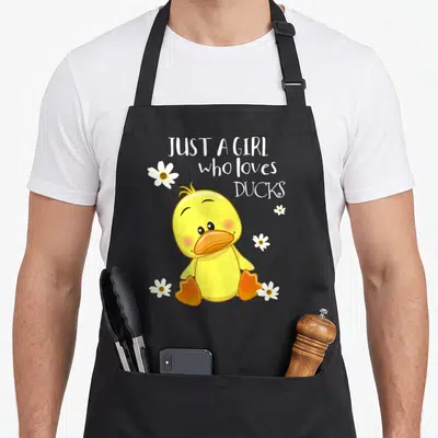 Owner duck apron - Yellow cartoon duck with orange feet surrounded by five white flowers and stacked text — shown on a black cotton canvas apron against a white background