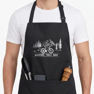 Trail bike apron - White line-art bicycle with mountains pine trees and text MOUNTAINS - TRAILS - BIKES — shown on a black cotton canvas apron against a white background