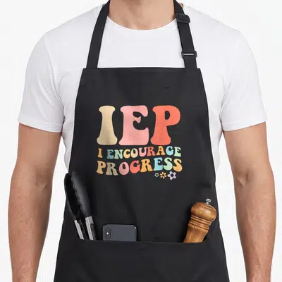 Iep teacher apron - Stacked IEP text in beige pink coral blue green and yellow with small flower cluster below — shown on a black cotton canvas apron against a white background