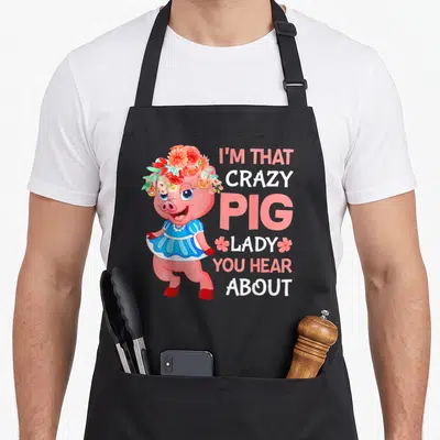 Funny hear crazy pig lady apron - Pink cartoon pig in blue dress and flower crown next to stacked text I'M THAT CRAZY — shown on a black cotton canvas apron against a white background