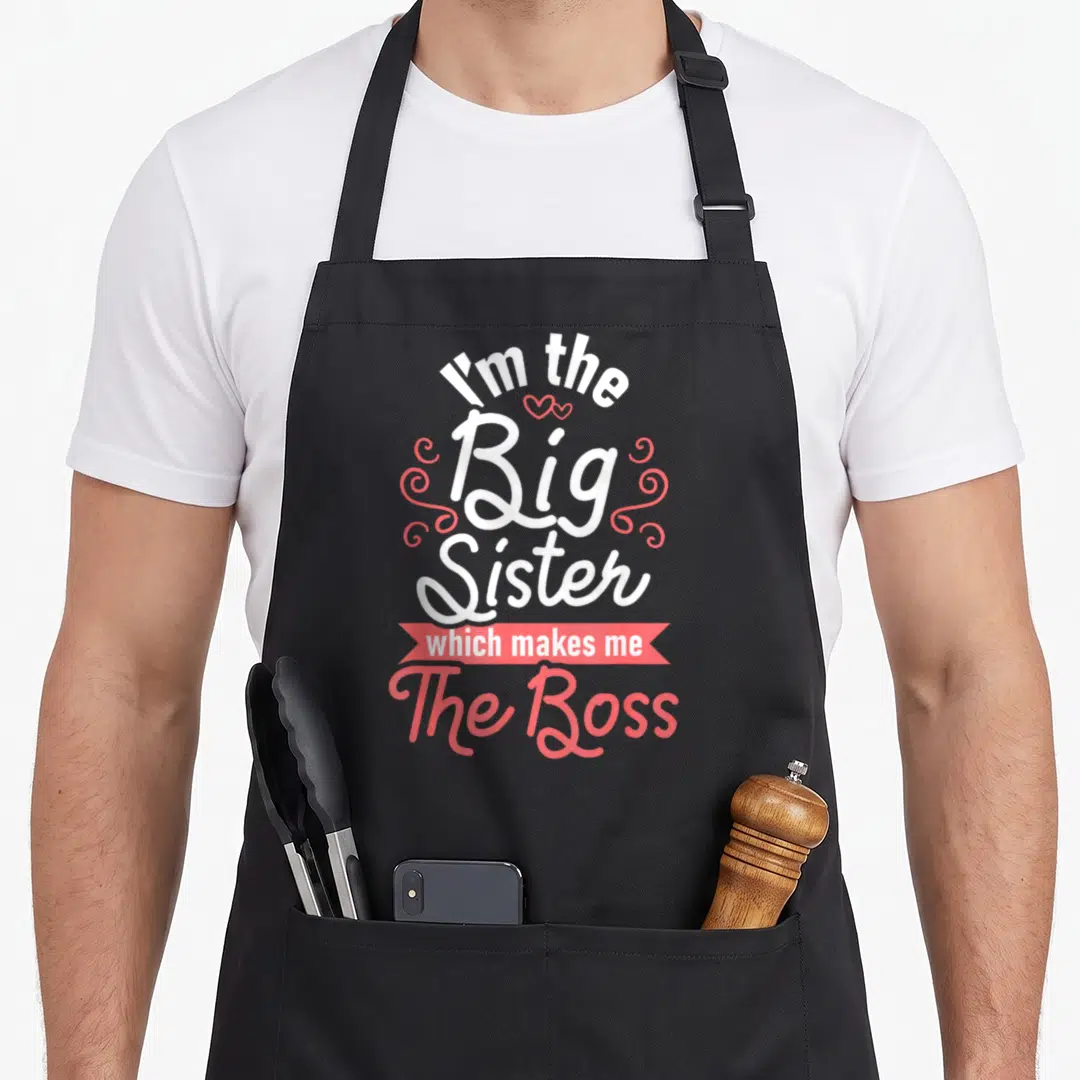 Sister boss apron - I'm the Big Sister which makes me The Boss text with pink swirls and two hearts — shown on a black cotton canvas apron against a white background