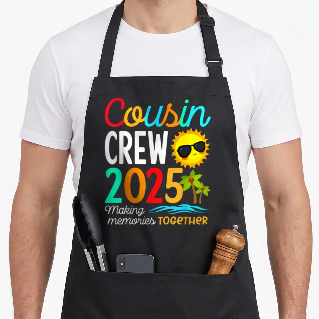 Summer vacation family aprons - Stacked colorful text Cousin CREW 2025 with cartoon sun sunglasses and two palm trees — shown on a black cotton canvas apron against a white background