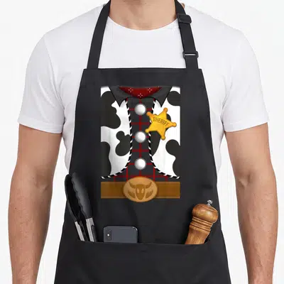 Woody halloween apron - Sheriff outfit with cow-print vest, red grid shirt, gold SHERIFF badge, and brown belt buckle — shown on a black cotton canvas apron against a white background