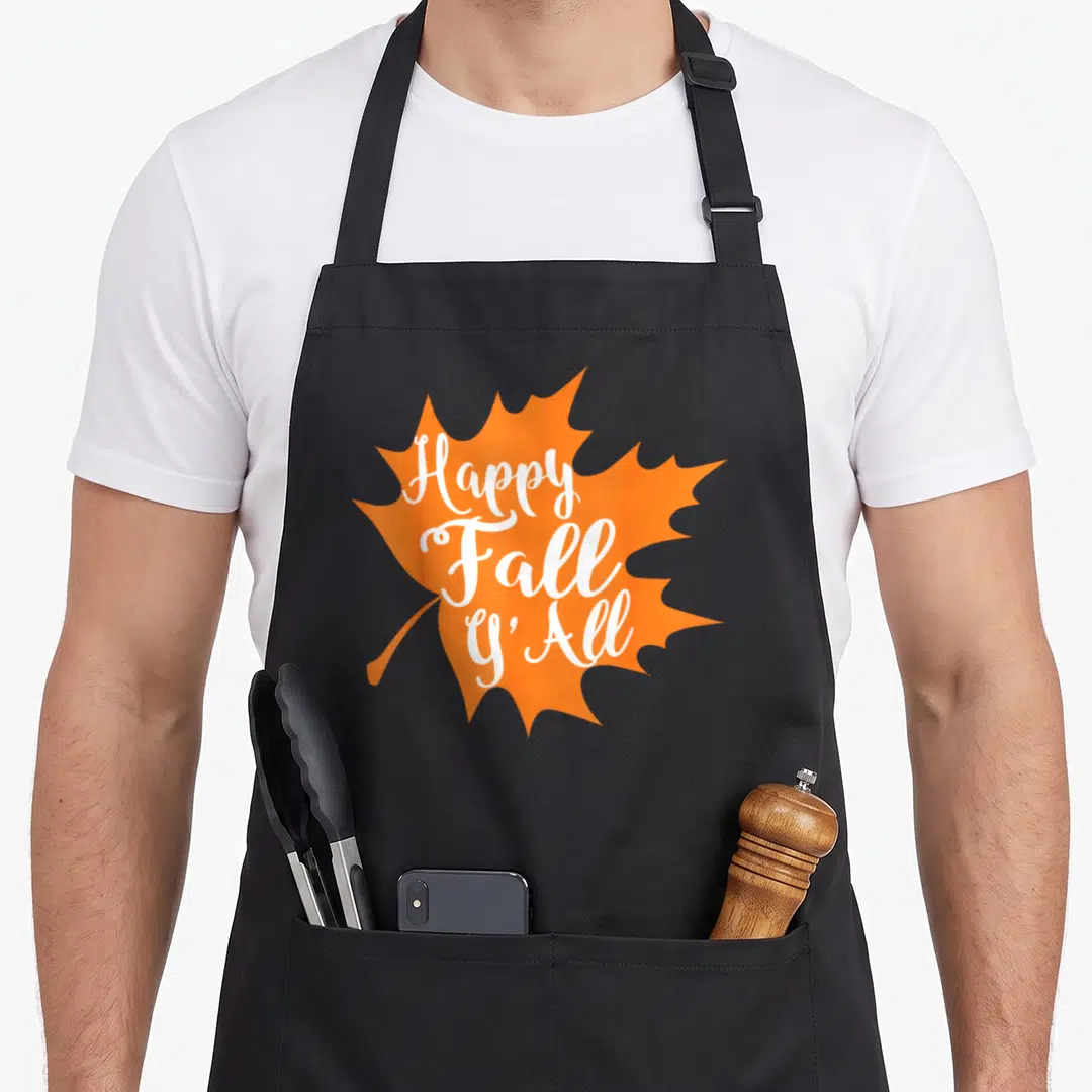 Yall cute fall aprons - Orange maple leaf with white script reading Happy Fall y'All — shown on a black cotton canvas apron against a white background