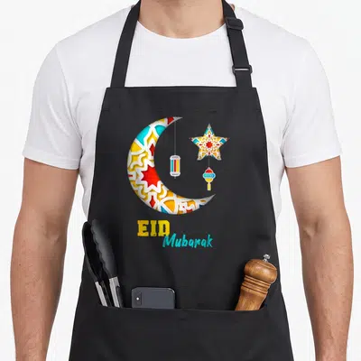 Alfitr eid apron - Patterned crescent moon star and two lanterns with EID Mubarak text — shown on a black cotton canvas apron against a white background