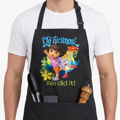 Dora explorer apron - Dora and Boots surrounded by tropical plants with ¡Lo hicimos! We did it! text — shown on a black cotton canvas apron against a white background