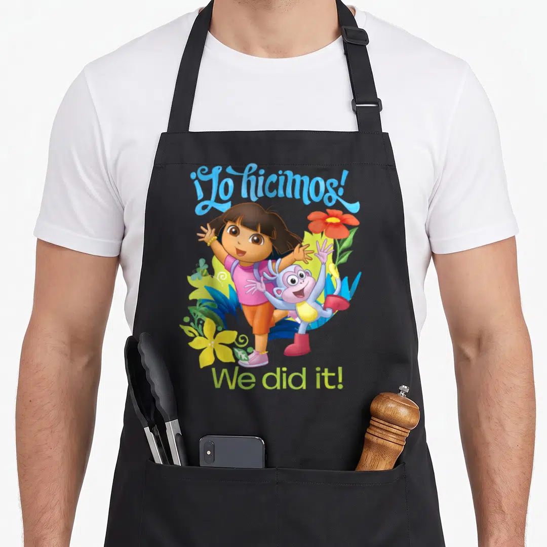 Dora explorer apron - Dora and Boots surrounded by tropical plants with ¡Lo hicimos! We did it! text — shown on a black cotton canvas apron against a white background