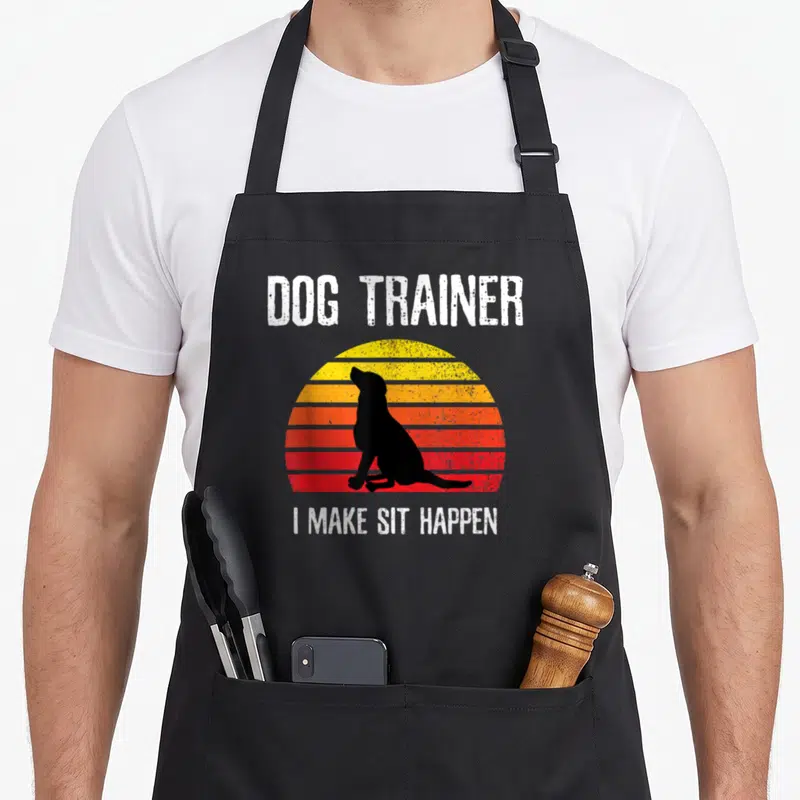 Command obedience dog training apron - Retro sun stripes and black dog silhouette with DOG TRAINER text — shown on a black cotton canvas apron against a white background