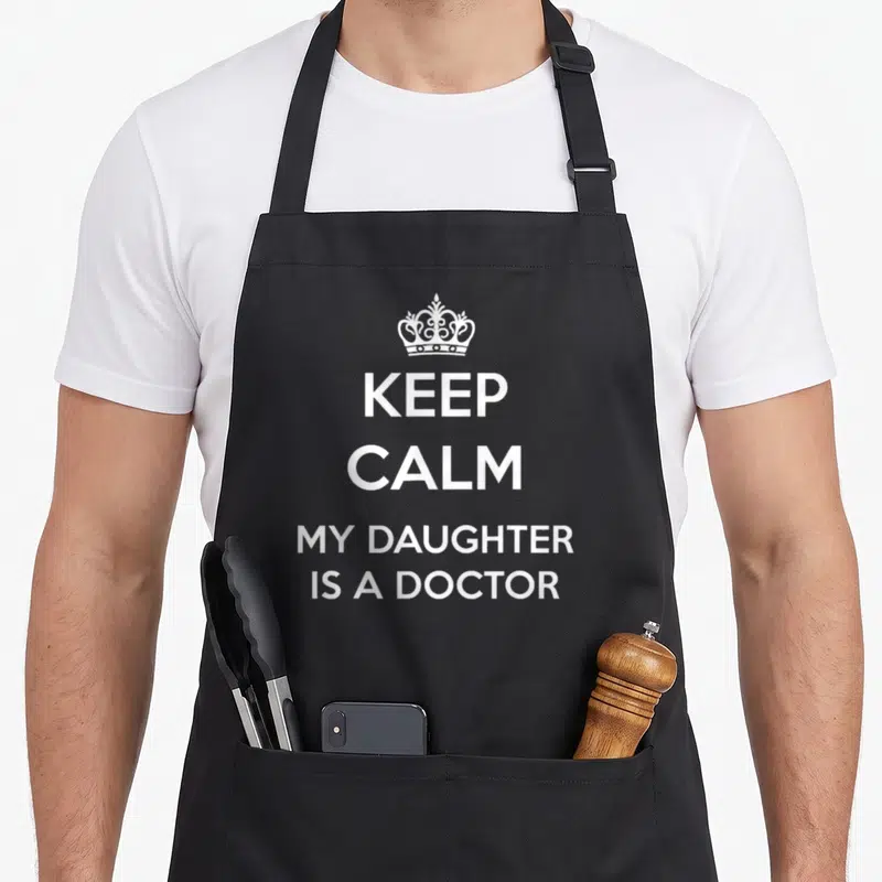 Daughter doctor with apron - White crown above four lines reading keep calm my daughter is a doctor — shown on a black cotton canvas apron against a white background