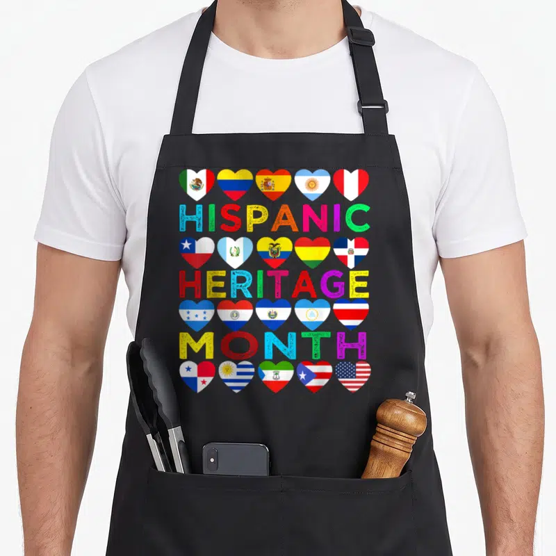 Latino country apron - Stacked HISPANIC HERITAGE MONTH text separated by rows of heart-shaped flags in red, yellow — shown on a black cotton canvas apron against a white background