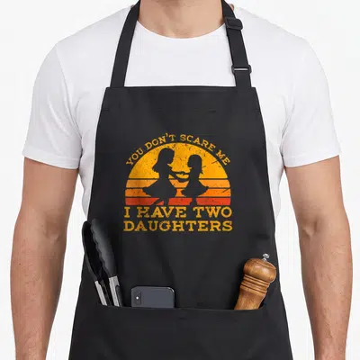 Cool scare father day apron - Retro orange sunset with two girl silhouettes and text You Don't Scare Me I Have Two — shown on a black cotton canvas apron against a white background