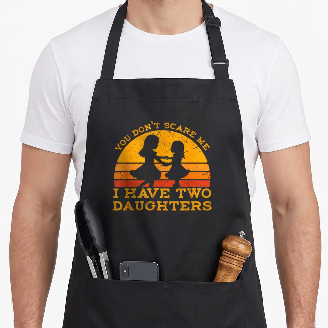 Cool scare father day apron - Retro orange sunset with two girl silhouettes and text You Don't Scare Me I Have Two — shown on a black cotton canvas apron against a white background
