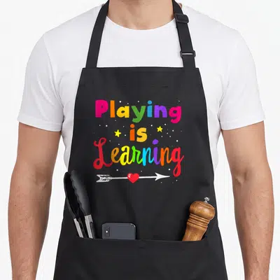 Childhood special aprons - Stacked colorful text Playing is Learning with two stars, dots, and a heart arrow below — shown on a black cotton canvas apron against a white background