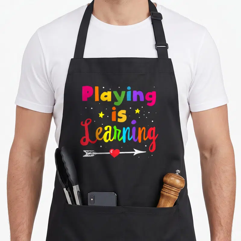 Childhood special aprons - Stacked colorful text Playing is Learning with two stars, dots, and a heart arrow below — shown on a black cotton canvas apron against a white background