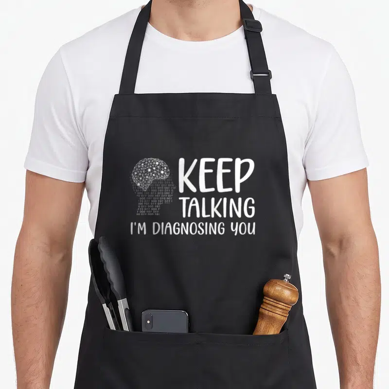 Graduation psychologist apron - White geometric brain overlapping binary code next to stacked text KEEP TALKING I'M — shown on a black cotton canvas apron against a white background