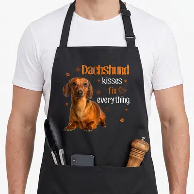 Kiss dachshund apron - Brown dachshund photo with orange flowers lips and text Dachshund kisses fix everything — shown on a black cotton canvas apron against a white background