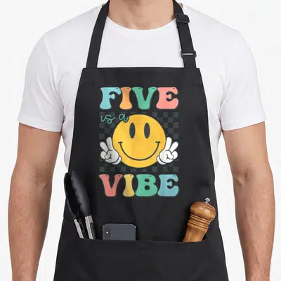 5th custom face apron - FIVE is a VIBE text with yellow smiley holding two peace signs on grey checkerboard — shown on a black cotton canvas apron against a white background