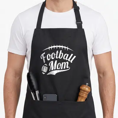 Proud sport football apron - White distressed Football Mom script with football icon and laces arch — shown on a black cotton canvas apron against a white background