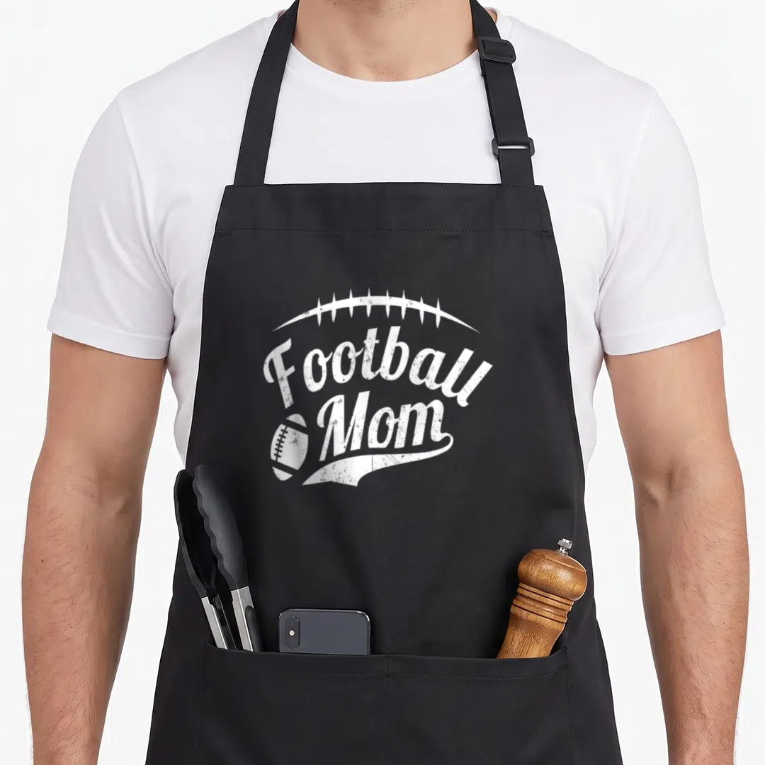 Proud sport football apron - White distressed Football Mom script with football icon and laces arch — shown on a black cotton canvas apron against a white background