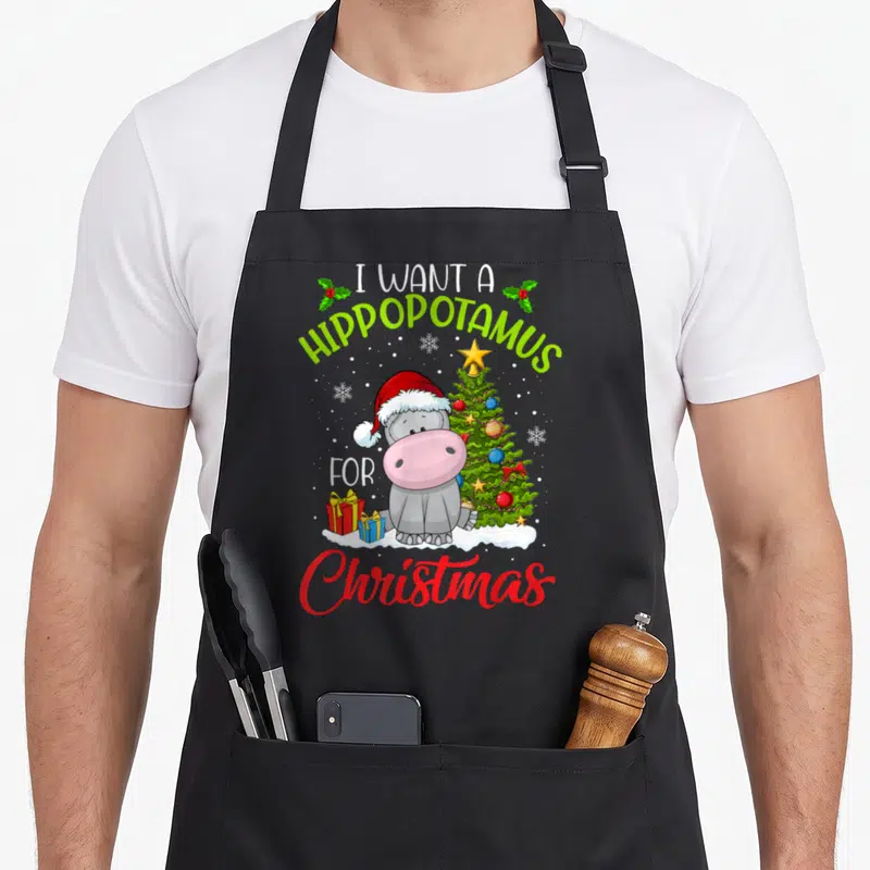 Hippopotamus christmas apron - Cartoon grey hippo in red Santa hat beside green Christmas tree and gifts on snow — shown on a black cotton canvas apron against a white background