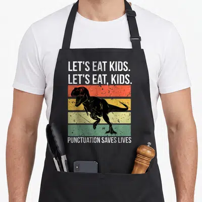 Save let's eat apron - T-Rex over red yellow green stripes with text LET'S EAT KIDS PUNCTUATION SAVES LIVES — shown on a black cotton canvas apron against a white background
