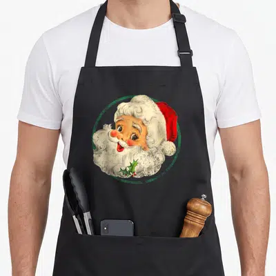 Claus old fashioned aprons - Retro Santa Claus face in green circle with holly sprig in beard — shown on a black cotton canvas apron against a white background