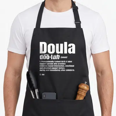 Noun doula apron - White typography defining Doula as a birth support person ending with 2. me — shown on a black cotton canvas apron against a white background