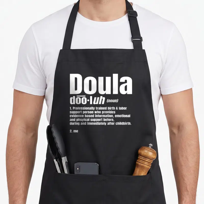 Noun doula apron - White typography defining Doula as a birth support person ending with 2. me — shown on a black cotton canvas apron against a white background