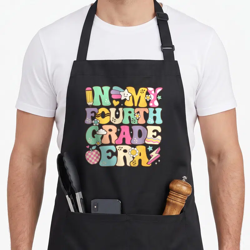 Era groovy grade apron - Stacked retro text IN MY FOURTH GRADE ERA with colorful pencil, apple, stars and flowers — shown on a black cotton canvas apron against a white background