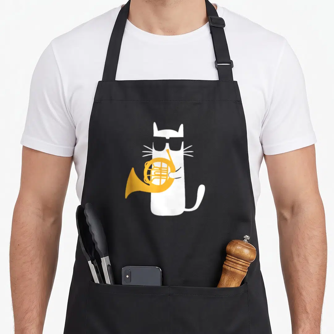 French cat apron - White cat with black sunglasses holding an orange french horn on a black bib — shown on a black cotton canvas apron against a white background