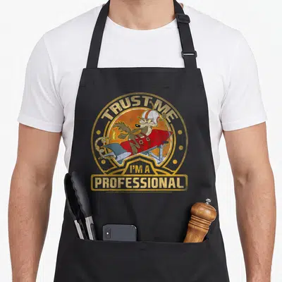 Wile professional aprons - Wile E. Coyote on red rocket inside yellow circle with gold TRUST ME I'M A PROFESSIONAL text — shown on a black cotton canvas apron against a white background