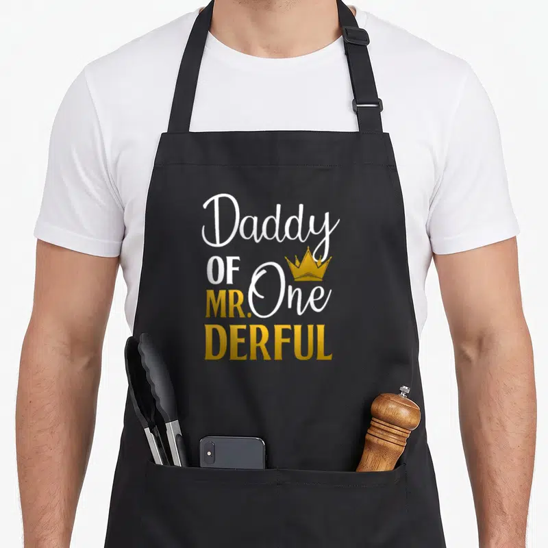 Daddy onederful father day apron - Stacked white and gold text reading Daddy OF MR One DERFUL with a gold crown — shown on a black cotton canvas apron against a white background