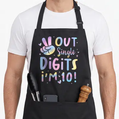 Peace birthday apron - Hand peace sign with OUT Single DiGiTS i'M 10! text in pastel gradient colors — shown on a black cotton canvas apron against a white background